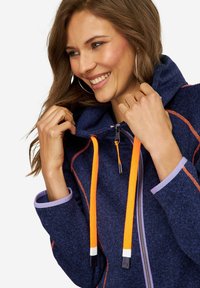 Veste à capuche zippée marine avec cordons de serrage orange, accents violets sur les poignets et un tissu texturé. Présente un col haut et un zip avant.