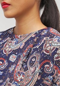 Marinblus med blommönster i paisley, mjukt tyg, rund halsringning och subtila veckdetaljer vid axeln.