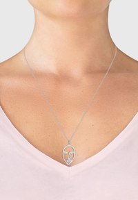 Silberne Halskette mit einem minimalistischen Gesichtdesign und einer stilisierten Handgeste, die an einer feinen Kette hängt und über einem hellrosa Oberteil getragen wird.
