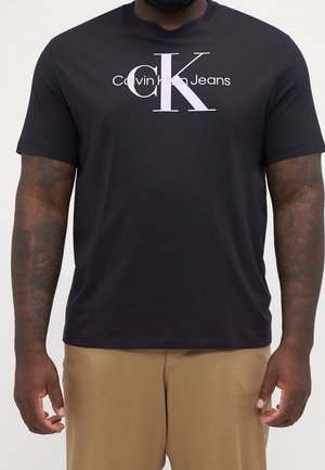 Homme portant un t-shirt noir Calvin Klein Jeans avec un grand logo blanc "cK", assorti d'un pantalon kaki, montrant ses avant-bras tatoués.