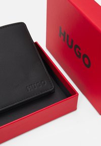 Black leather wallet with embossed "HUGO" logo, placed inside a red box with a black interior. Edges are neatly stitched.