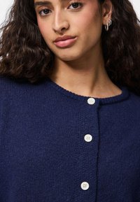 Navy blue knitted cardigan with a round neckline, featuring five white buttons down the front and a textured fabric.