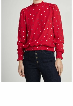 Blusa rossa a pois con colletto arruffato e maniche lunghe, realizzata in tessuto testurizzato, abbinata a jeans scuri a vita alta con dettagli a bottoni.