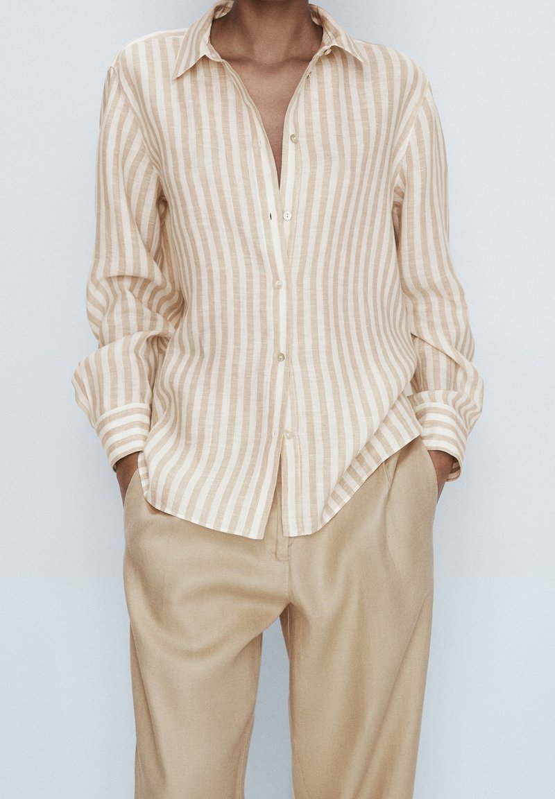 Personne portant une chemise à rayures verticales beige et blanche à boutons, manches retroussées, et un pantalon beige clair avec les mains dans les poches.