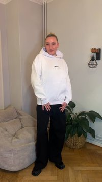 Sweatshirt branca com capuz e logótipo preto, calças largas pretas, sapatos pretos; ambiente interior com um sofá bege texturizado e uma planta verde próxima.