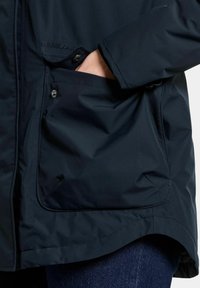 Marin vattentät jacka med en slät yta, som har en synlig ficka med tryckknappsstängning och märkesdetaljer på framsidan.