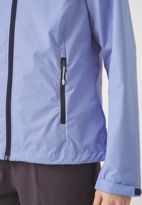 Veste imperméable violet clair avec une fermeture éclair noire et une poche latérale. Elle présente une texture lisse et des poignets ajustables pour un ajustement sécurisé.