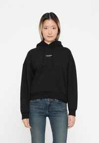 Svart kort hoodie med snorhette og frontlomme. Har hvit tekstlogo, avslappet passform, lange ermer og myk stofftekstur.