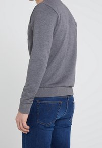 Personne portant un pull à manches longues gris et un jean en denim bleu, debout contre un fond clair uni, de profil.