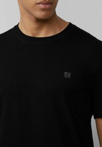 Schwarzes Baumwoll-T-Shirt mit Rundhalsausschnitt, kurzen Ärmeln und einem kleinen schwarzen Logopatch auf der linken Brustseite.