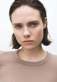 Porträt einer Person mit kurzen, dunklen Haaren, die ein hellbraunes Hemd trägt. Zu den Merkmalen gehören helle Haut und ein neutraler Gesichtsausdruck.