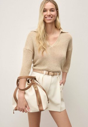 Usmievavá žena s dlhými blond vlasmi, oblečená v béžovom pletenom svetri a krémových kraťasoch s opaskom, drží veľkú bielu a svetlohnedú koženú kabelku.