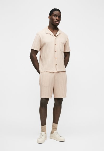 Homme debout portant une chemise à manches courtes boutonnée texturée beige clair avec un short assorti, des chaussettes beiges et des baskets blanches sur un fond uni.