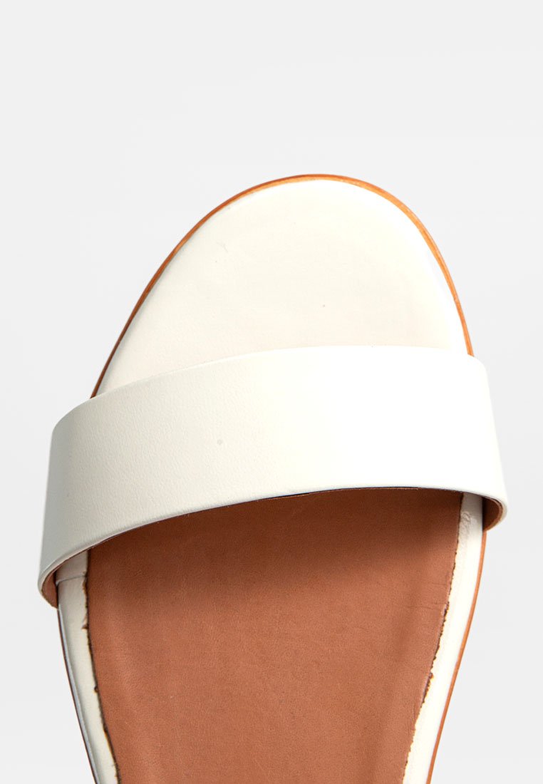 Sandalia deslizante blanca con una banda ancha y una suela marrón. Textura de cuero suave, diseño de punta redonda, estilo minimalista, sin hardware visible.