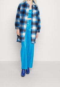 Giacca camicia a quadri blu e nera con accenti bianchi, indossata sopra una maglia blu lucida e pantaloni di raso abbinati, completata da calzature blu.