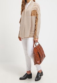 Beżowy kardigan w wzory z frędzlami, biała koszula, białe dżinsy i czarne buty wsuwane. Brązowa skórzana torebka z dwoma przednimi kieszeniami.
