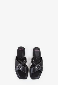Sorte faux læder sandaler med firkantet tå, der har krydsede stropper og sølvspænde detaljer. Indersålen har en glat overflade.
