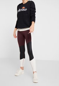 Sudadera negra con logo, superpuesta sobre una blusa blanca con lunares. Leggings granates y negros con diseño en bloques de color. Zapatillas deportivas blancas.