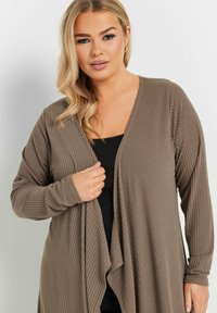 Brun, ribbad cardigan med långa ärmar och öppen front, lager på lager över en svart topp, med en mjuk textur och avslappnad passform.