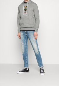 Sudadera gris con estampado gráfico, bolsillo frontal y capucha con cordón, combinada con jeans azules deslavados y zapatillas negras.