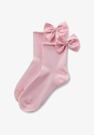 Para różowych skarpetek do kostek, z których każda ozdobiona jest dużą różową satynową kokardą przyszytą blisko ściągacza, ułożonych płasko na białym tle.