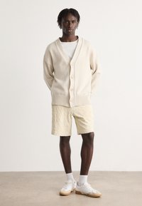 Beige knitted cardigan with V-neck, ribbed hem, and beige buttons, worn over a white T-shirt. Light yellow textured shorts and white sneakers.