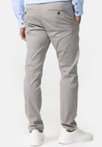 Man in een lichtgrijze slim-fit broek, witte sneakers en een lichtblauw shirt, van achteren weergegeven met zijn handen in de zakken.