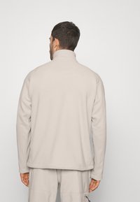 Lys beige fleecejakke med høj krave, flade sømme og lange ærmer. Glat tekstur, afslappet design, velegnet til lag-på-lag.