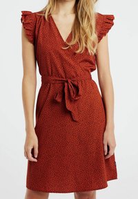 Röd klänning med svart prickmönster, v-ringning, korta volangärmar och ett knytbälte i midjan. Slät tyg med en figursydd form.