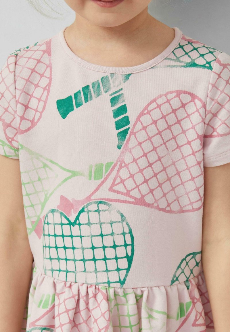Kind trägt ein blassrosa Kleid mit kurzen Ärmeln, das grüne und pinke abstrakte Herz- und Netzmuster zeigt.