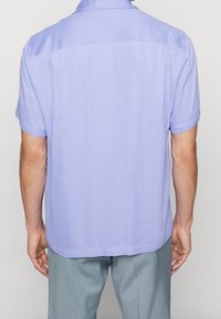 Uomo che indossa una camicia lavanda a maniche corte e pantaloni grigi chiari, visto da dietro su uno sfondo neutro.