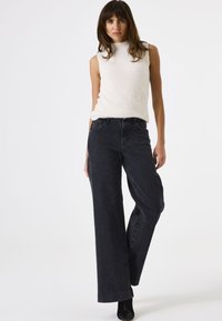 Vrouw die een mouwloos wit, pluizig topje draagt en wijde zwarte jeans met hoge taille en kleine witte vlekjes, staand tegen een effen lichte achtergrond.