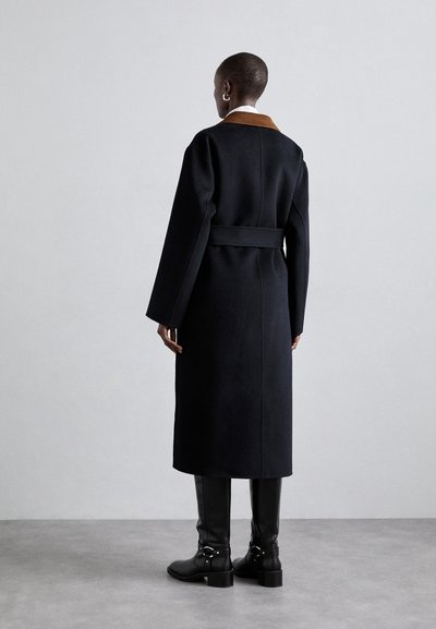 Langer schwarzer Wollmantel mit kontrastierendem braunem Kragen, tailliertem Schnitt und gerader Silhouette. Schwarze knielange Lederstiefel mit Schnallenakzenten.