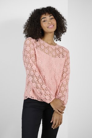 Haut en dentelle rose à manches longues, présentant des motifs floraux et un col festonné. Associé à un pantalon sombre.