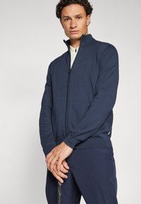 Sweater azul-marinho com fecho zip em malha texturizada. Apresenta um colarinho alto, punhos e bainha canelados, com uma pequena etiqueta da marca no lado esquerdo.