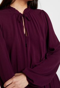 Lila klänning med en volangkrage, nyckelhålsöppning och långa ärmar med rynkade manschetter, tillverkad av mjukt och slätt tyg.