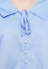 Jasnoniebieska koszula z karbowanym kołnierzem, wiązanym przodem oraz pięcioma okrągłymi perłowymi guzikami biegnącymi wzdłuż środka. Gładka faktura materiału.