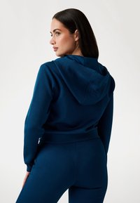 Felpa blu scuro con cappuccio e tessuto texture, caratterizzata da un design cropped. Abbinata a pantaloni blu scuro aderenti, che mettono in evidenza un aspetto uniforme.