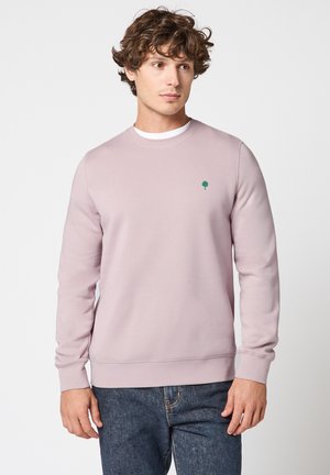 Homme aux cheveux bouclés portant un sweat-shirt mauve clair avec un petit logo d'arbre vert sur la poitrine et un jean en denim bleu.