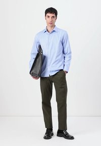 Gaiši zils garām piedurknēm krekls, olīvu zaļas bikses, melni ādas apavi un melna rokassoma. Modelis stāv pretī vienkāršai baltai fona.