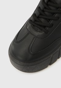 Fekete bőr sportcipő robusztus szürke talppal, kerek fűzőkkel és a felsőrészén látható varrási részletekkel, amelyek textúrát adnak az összképhez.