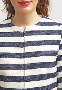 Pruhovaná bunda z bílého a námořnicky modrého texturovaného materiálu, s zipem a kulatým výstřihem. Detaily se lesklým kováním zipu.