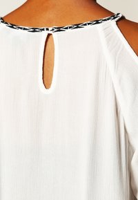 Vit topp med ett nyckelhål i ryggen, prydd med en svart mönstrad kant kring halslinjen, mjukt texturerat tyg.