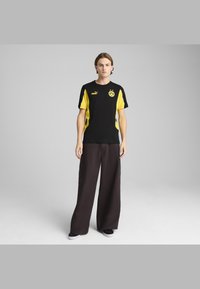Černé a žluté sportovní tričko s logem, krátké rukávy, spárované s tmavými širokými kalhotami a černými botami. Hladká tkanina, uvolněný střih.