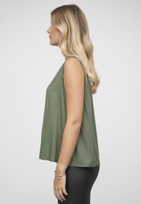 Ermeløs topp i olivengrønn, laget av mykt stoff med en løs passform. Rundt skjørt og minimale sømmer, kombinert med svarte leggings.