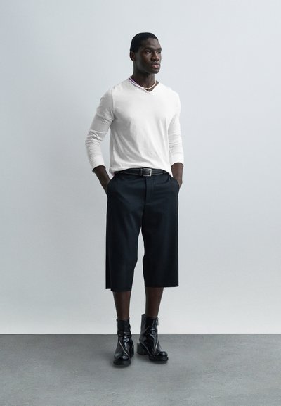 Άνδρας στέκεται με τα χέρια στις τσέπες, φορώντας λευκό πουκάμισο με μακριά μανίκια, μαύρο παντελόνι cropped, μαύρες μπότες με τακούνι και κολιέ με χάντρες σε γκρι φόντο.