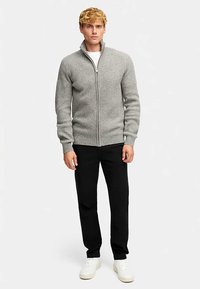 Šedý žebrovaný svetr na zip s vysokým límcem, kombinovaný s černými kalhotami a bílými teniskami, ukazující přiléhavý střih a texturovanou tkaninu.