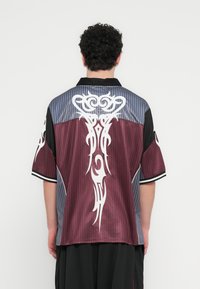 Κοντομάνικο πουκάμισο με μπορντό σώμα, γκρι λεπτομέρειες και λευκά φυλετικά σχέδια στην πλάτη. Κατασκευασμένο από λείο, ελαφρύ ύφασμα.