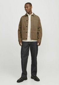 Chaqueta de algodón marrón con bolsillos frontales, cierre de botones y cuello puntiagudo, usada sobre una camisa a rayas y jeans de mezclilla oscuros. Zapatos negros.