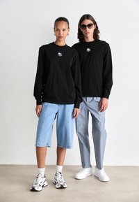 Zwei Models tragen schwarze Langarmhemden von Lacoste; die Frau in hellblauen Denim-Shorts und Turnschuhen, der Mann in hellblauen gestreiften Hosen und weißen Sneakers.